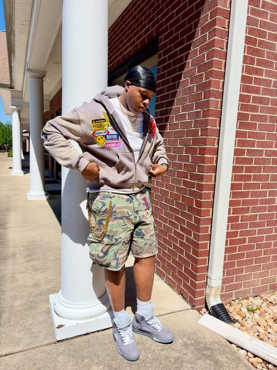 Customer wearing the Ash K1 Zip Up in washed taupe with camo cargo shorts and grey sneakers, leaning against a column outside a brick storefront.