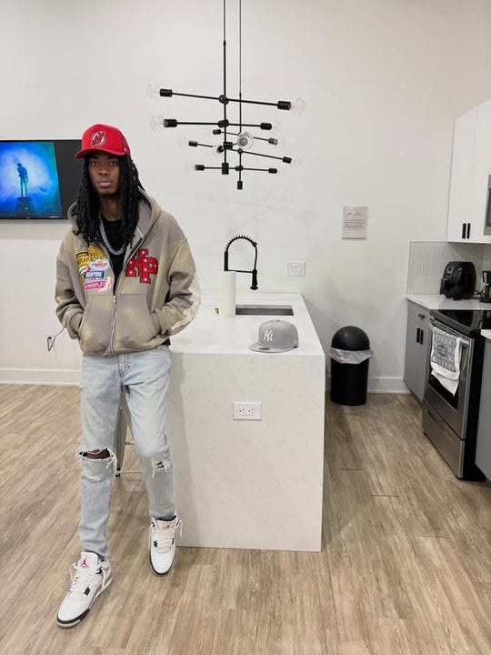 Customer wearing the Ash K1 Zip Up in washed taupe with light-wash ripped jeans and a red cap, standing in a kitchen.
