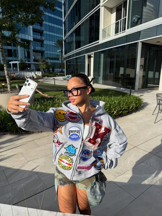 Customer wearing the grey Patchy Zip hoodie zipped over camo jorts, taking a phone selfie in a sunlit high-rise courtyard.
