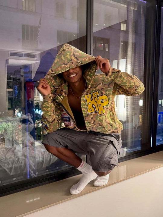 customer in a camo kings play zip up, hood up, by a window at night